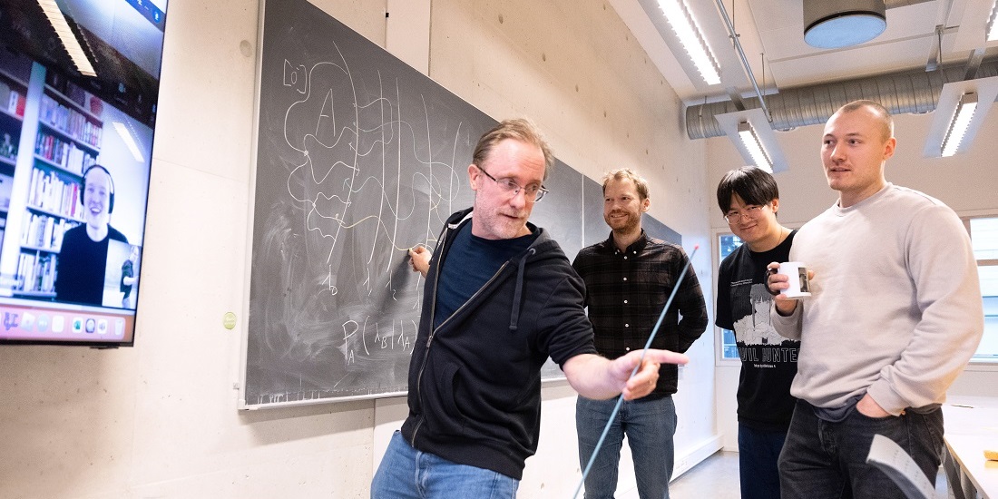 4 men standing in front of a blackboard. One is pointing and drawing on the blackboard