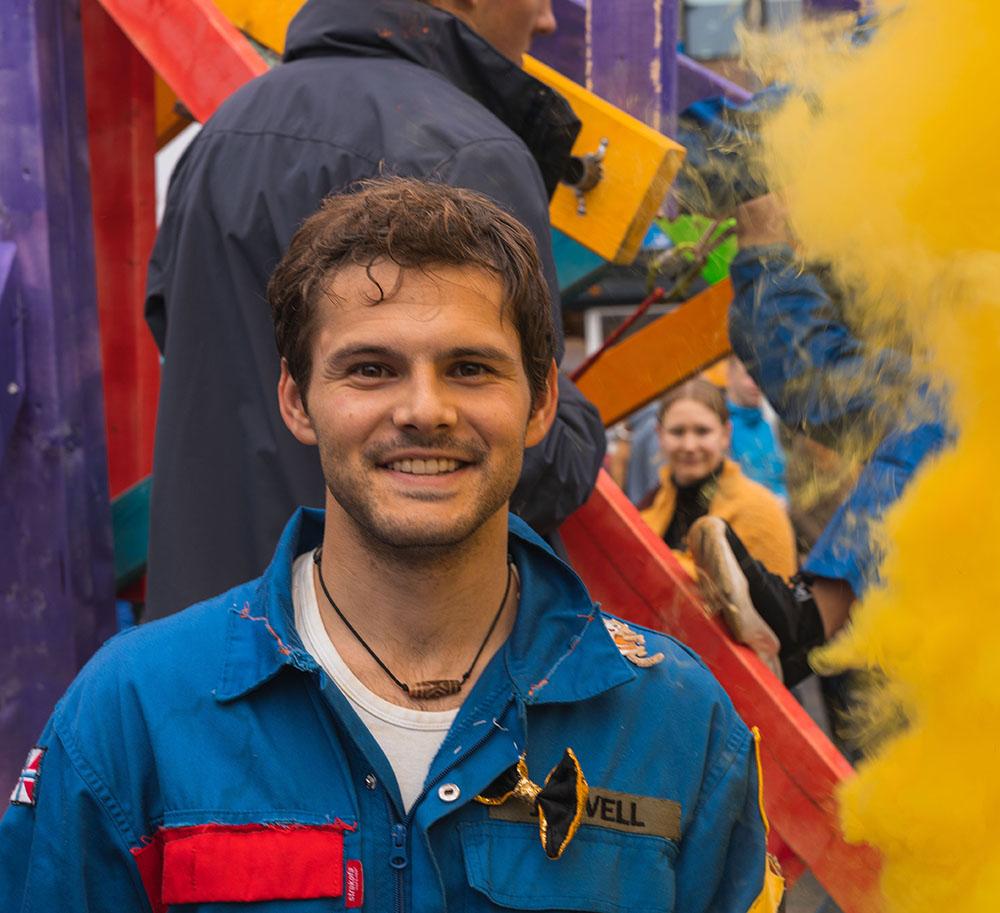 Male with brown hair and big smile wearing blue suit stands in front of a colourful background,