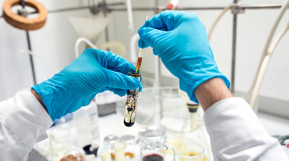 A pair of hands wearing blue rubber gloves stirs in a test tube and you see black liquid dissolving in clear fluid. Background is filled with various chemistry equipment. Photo