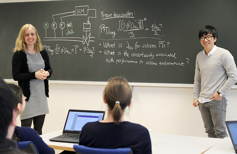 A teacher and a student in a classroom standing at a blackboard while standing in front of other students. Photo.