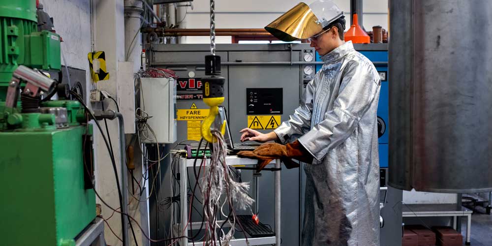 A student stands with shiny metal protective gear and a gold helmet in a lab. Photo. 