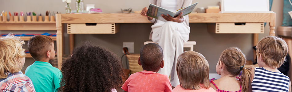 A group of children sit on the floor facing an adult who is reading a book aloud at the front of the room. The setting appears to be a classroom with shelves and art supplies in the background. Photo