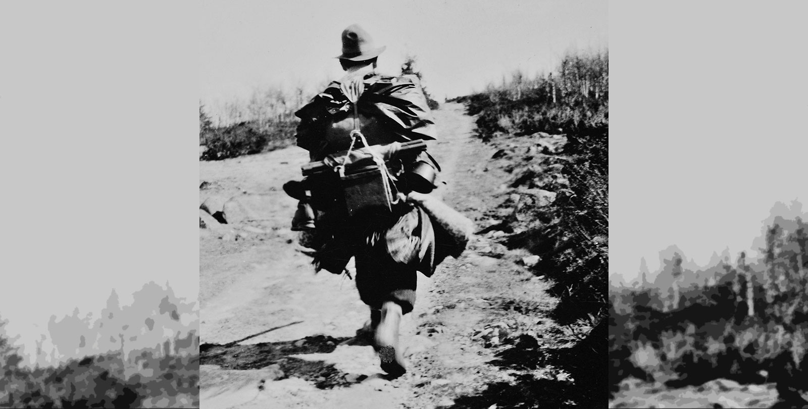 Historic black-and-white photograph showing a person walking barefoot along a dirt road. The person is carrying several items attached to their body and in their hands. The road is surrounded by vegetation and sloping terrain on both sides. Photo