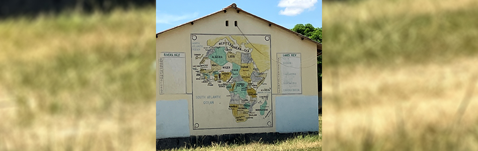 A light-colored brick wall with a large painted map of Africa. The map shows countries in different colors and includes the names of rivers and lakes written on the sides. The photo is taken outdoors with grass in the foreground and trees in the background. Photo
