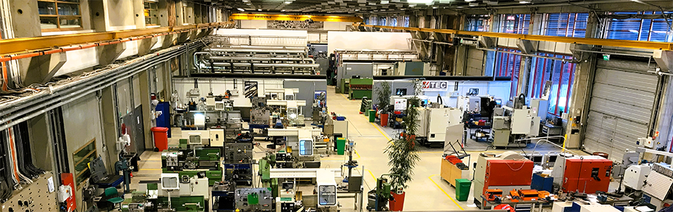 Overview image of a large industrial hall with machinery, production equipment, and workstations arranged in rows across the factory floor. Photo