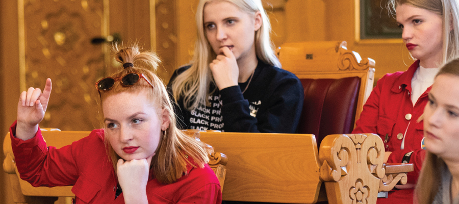 Several students are sitting in a parliamentary chamber with benches. One student raises a hand while others listen. Photo.