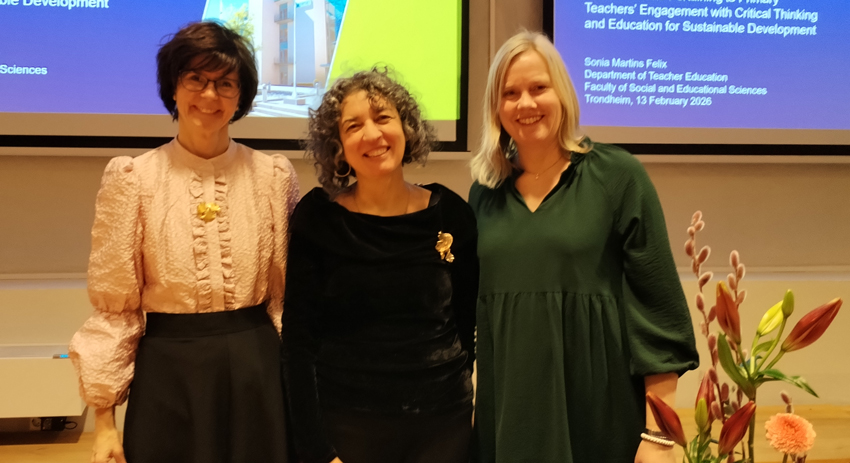 Three people stand side by side in front of a screen in a lecture hall. The screen displays a presentation about teacher education and sustainable development. The people are wearing clothes in different colors, and on the right side of the image there is a bouquet of flowers in a vase. Photo