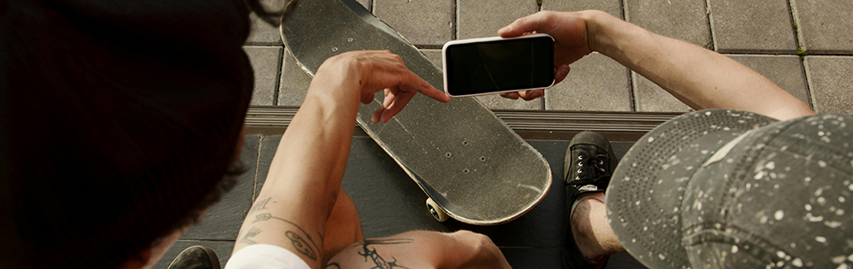 Two people are sitting outside on a curb with a skateboard between them. One is holding a smartphone while the other points at the screen. Photo