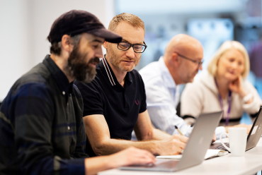 Four people are sitting at a table in a meeting room, with laptops in front of them. They appear to be engaged in a discussion or work meeting. There is also a white coffee cup on the table. Photo