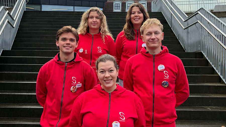 Five people are standing together in front of an outdoor staircase. All of them are wearing red hoodies with a white logo on the chest and name tags attached to the garment. The group is standing in two rows, with three people in front and two behind. Metal railings on both sides of the staircase are visible in the background. Photo.