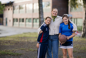An adult and two children stand together outdoors in front of a school building. The child on the right is holding a basketball, and the three are standing close as a group. Photo.
