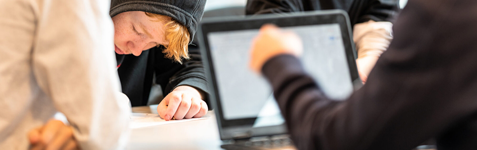 Three people are collaborating at a table. One person wearing a hoodie is looking down at a sheet of paper, while another is pointing at a laptop. A third person is partially visible in the foreground. Foto
