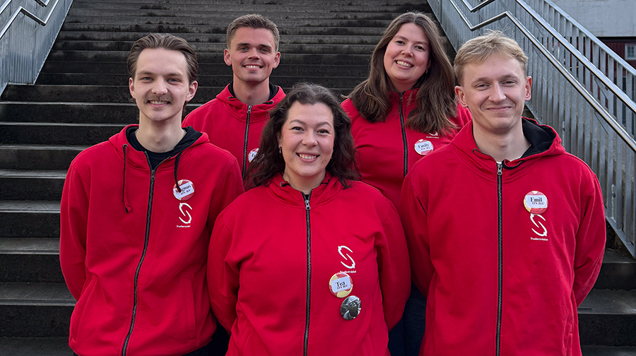 Five people are standing together in front of an outdoor staircase. All of them are wearing red hoodies with a white logo on the chest and name tags attached to the garment. The group is standing in two rows, with three people in front and two behind. Metal railings on both sides of the staircase are visible in the background. Photo.
