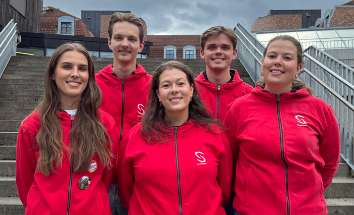 Five people are standing in front of an outdoor staircase, all wearing red hoodies with a white logo on the left chest. Behind them are brick buildings and a gray, overcast sky. Photo.