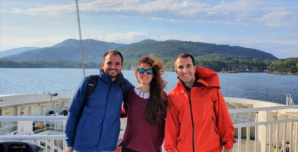 Three phd students staning on a ferry, with wind park in the background
