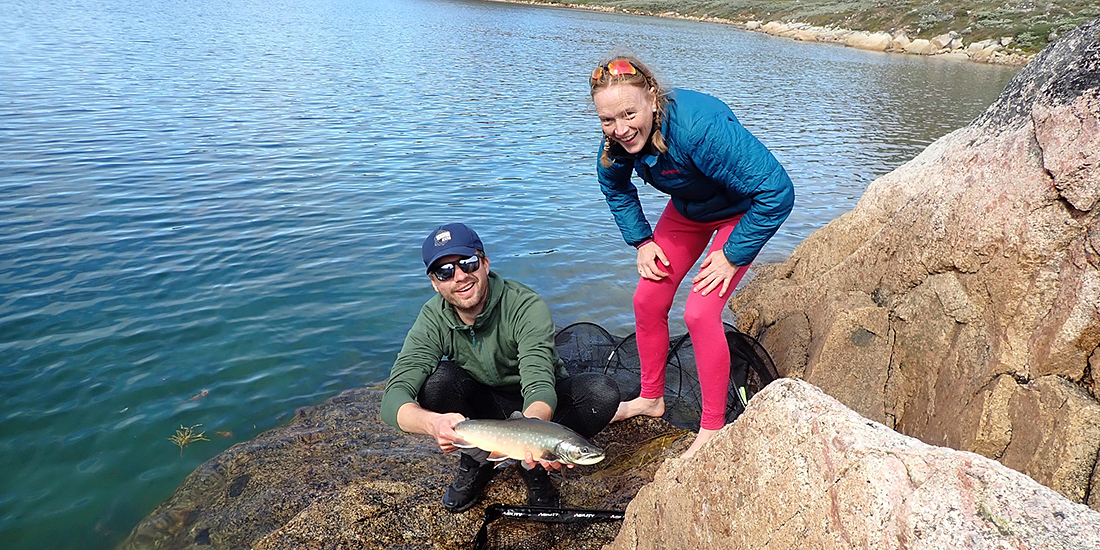 A man and a woman standing close to the ocean. The man is holding an arctic char, smiling and looking into the camera