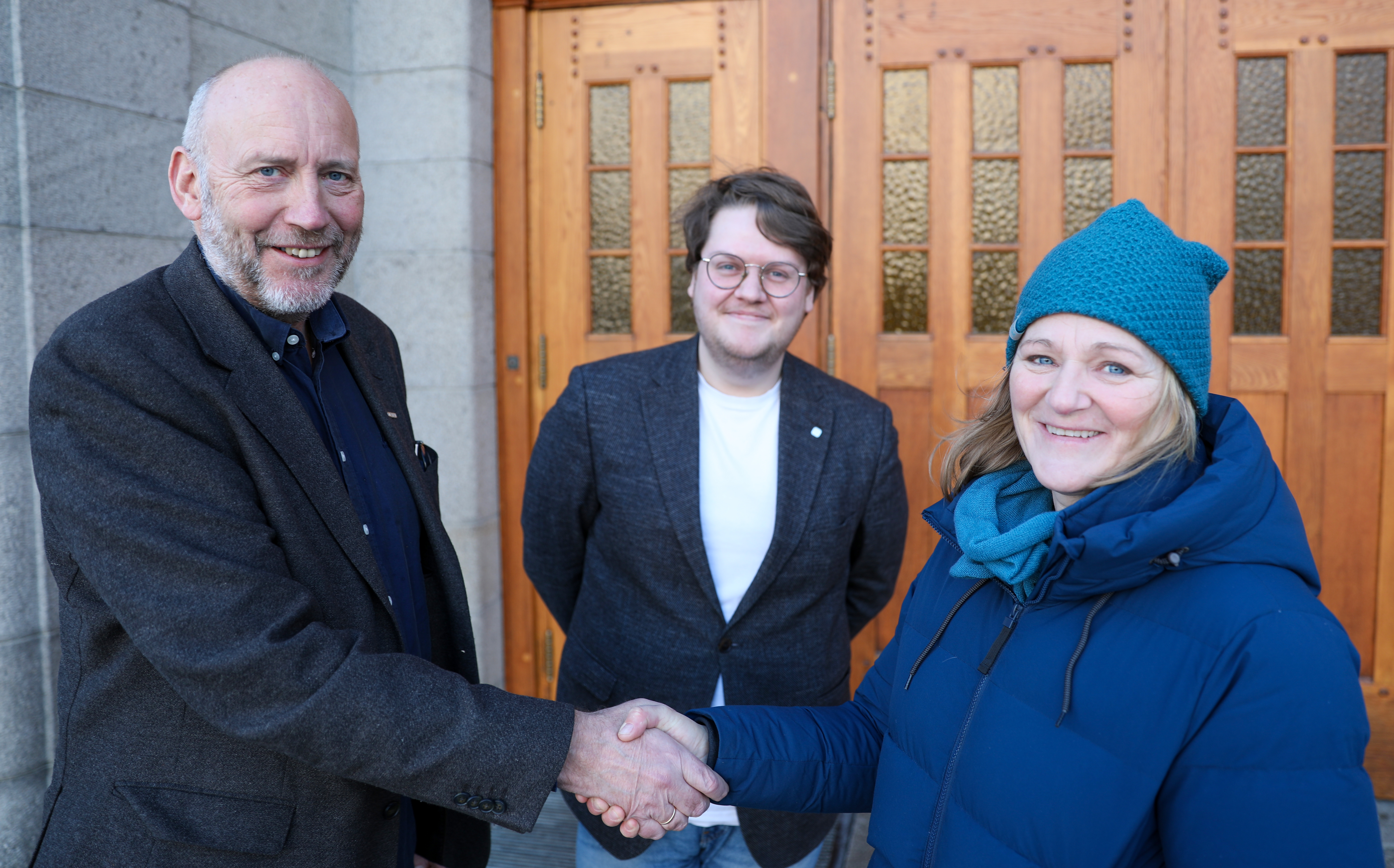 Rector Tor Grande and Katrin Glatz Brubakk shakes hands, and smiles at the camera. Behind them, Morten Eidsvaag Althe stands, smiling.