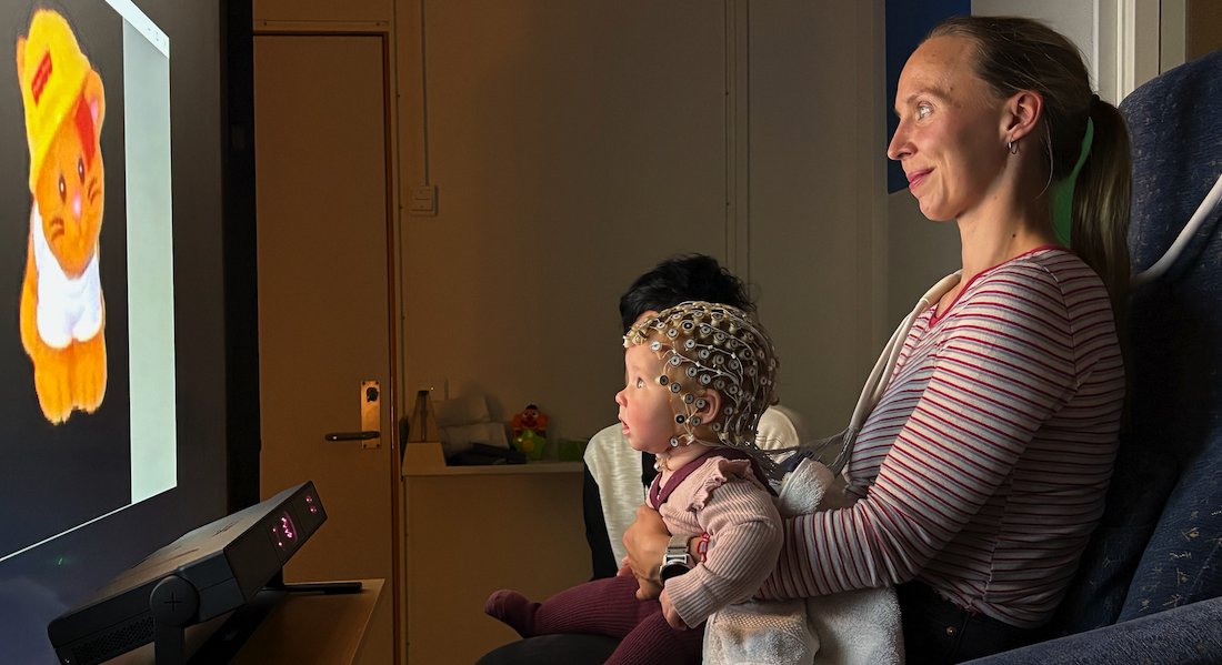 Mother and baby wearing cap that can measure brain activity in front of large screen