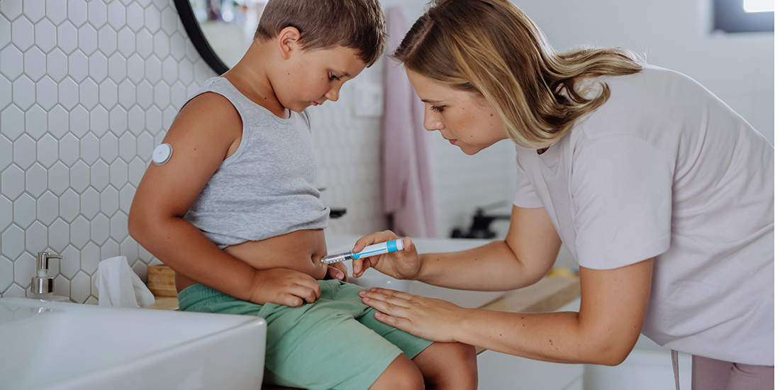 A young boy is sitting on a counter in a bathroom. A woman is giving him an injection with insulin