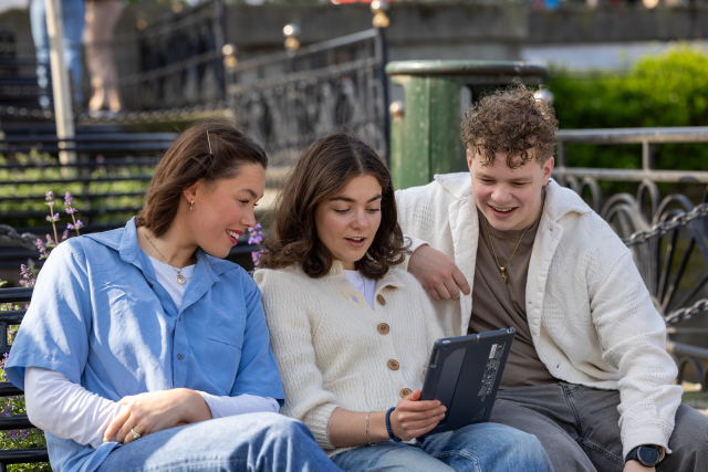 Three students sitting together on a bench looking at an ipad. 