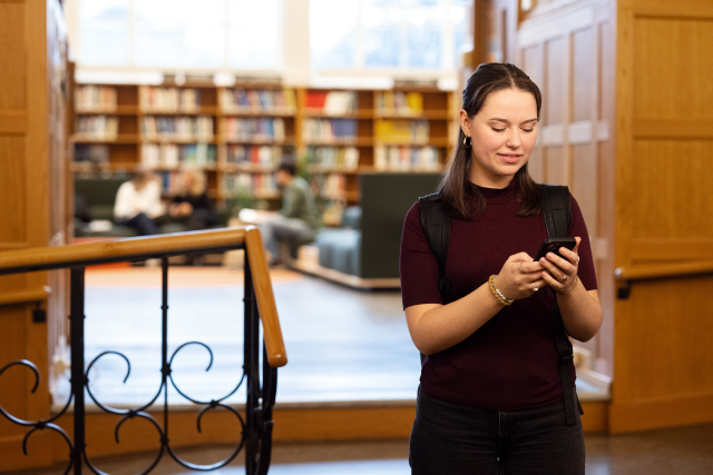 A girl standing infront of the library looking down at her phone. 