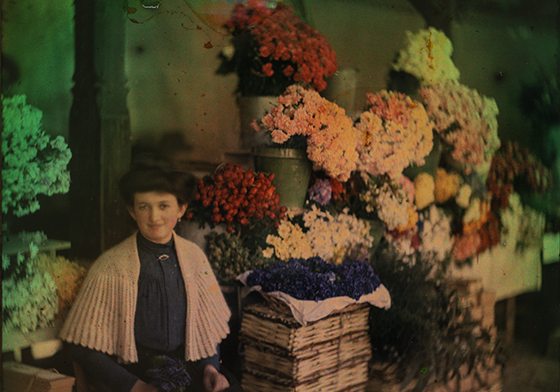 Woman with vintage hairdo and light-colured shawl sits in front of a colorful display of flowers.