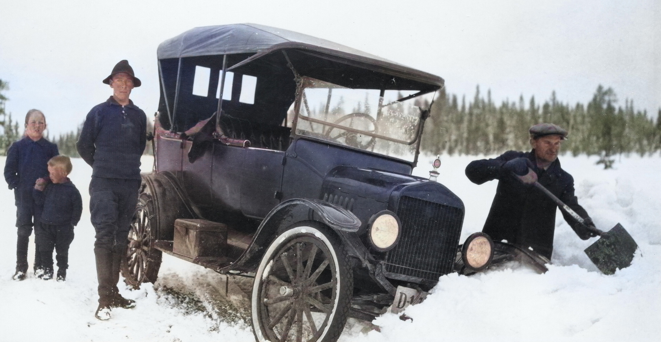 A car in the ditch. A man is clearing snow by the road, while another man and two boys are watching. Photo