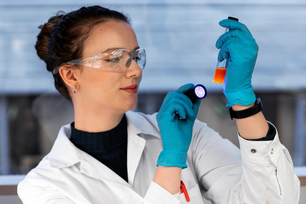 A female student in white lab coat and protective glassed is directing a flash light towards a fluroscent test tube. Photo. 
