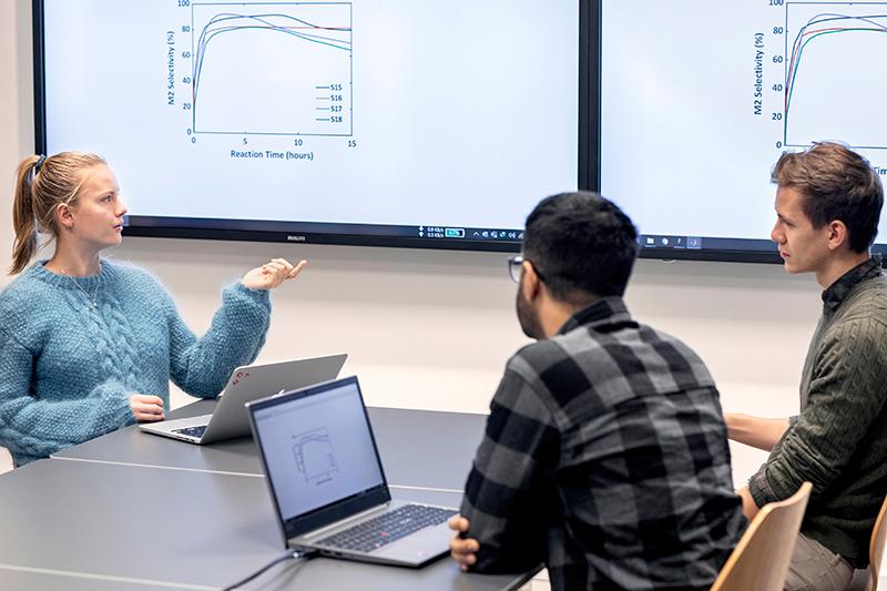 Students working together in front of a big screen displaying various graphs. Photo