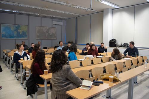 A group of approximately twenty adults engage in discussion while seated in a room with rows of tables.