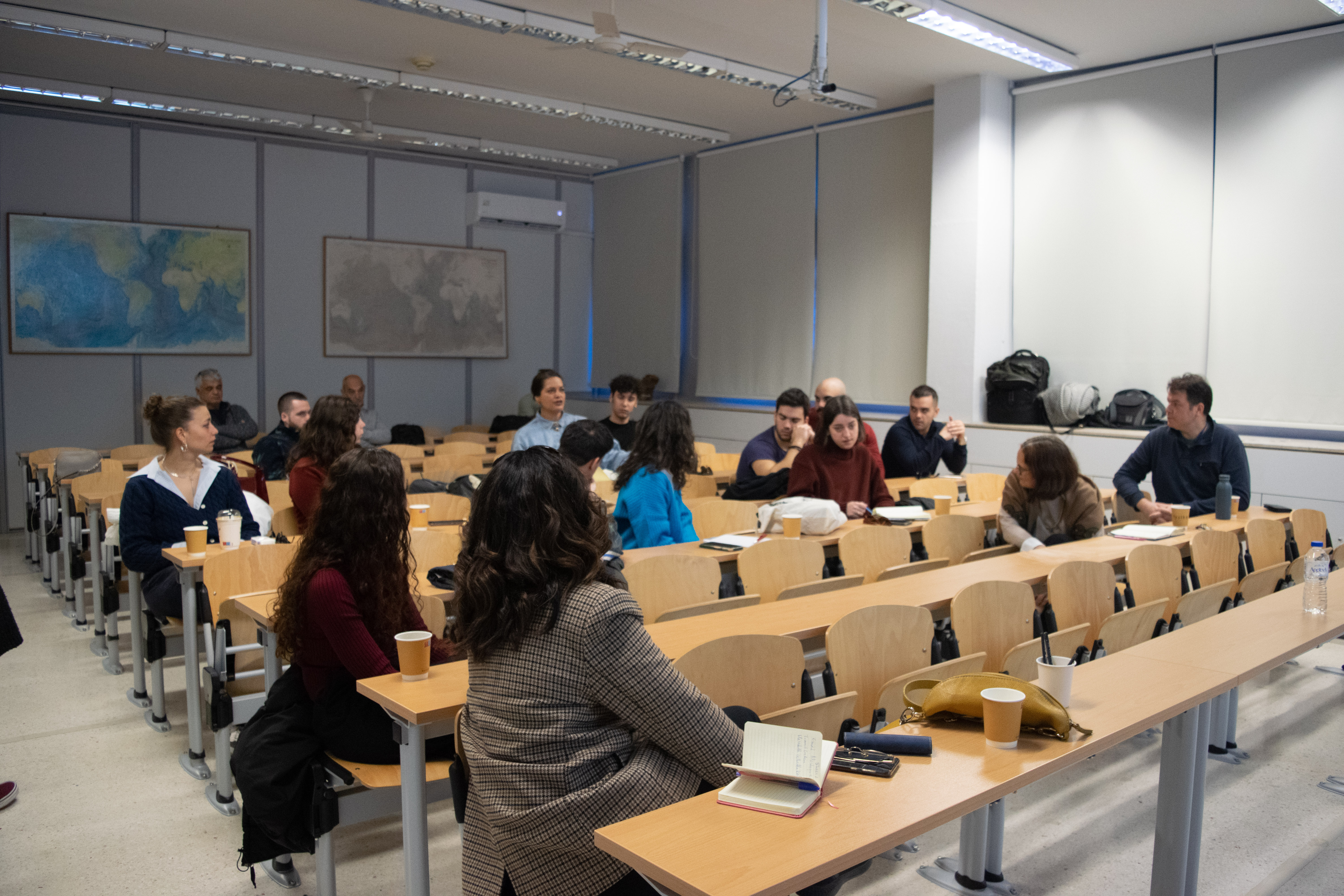 A group of approximately twenty adults engage in discussion while seated in a room with rows of tables.