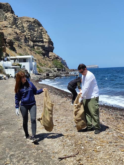 Three adults pick up items on a sandy beach in front of cliffs and blue sky and put them into bags.