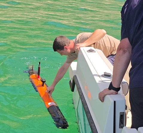 A researcher lowers marine robotic monitoring equipment into a turquoise blue sea.