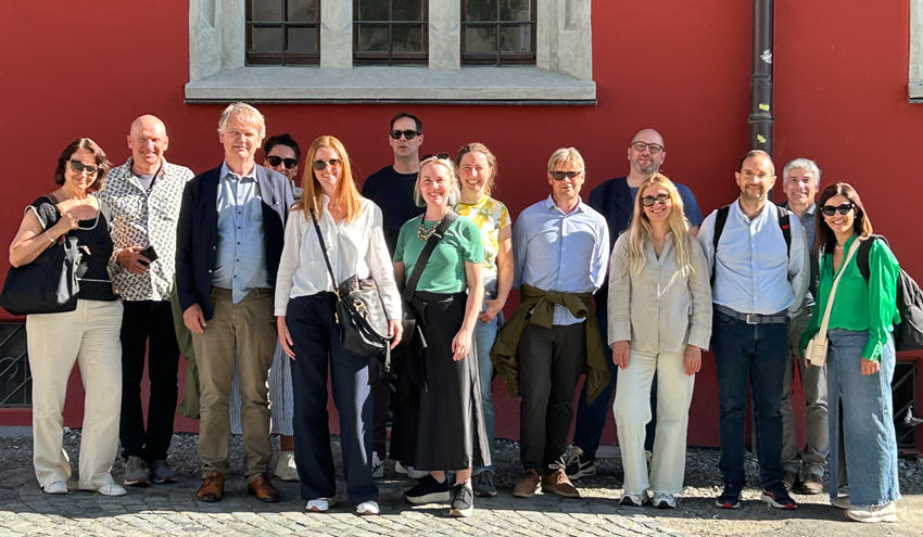 A group of people standing together outdoors in front of a building with a red wall and two windows. They are standing on cobblestone in sunlight, and several are carrying bags or jackets. Photo
