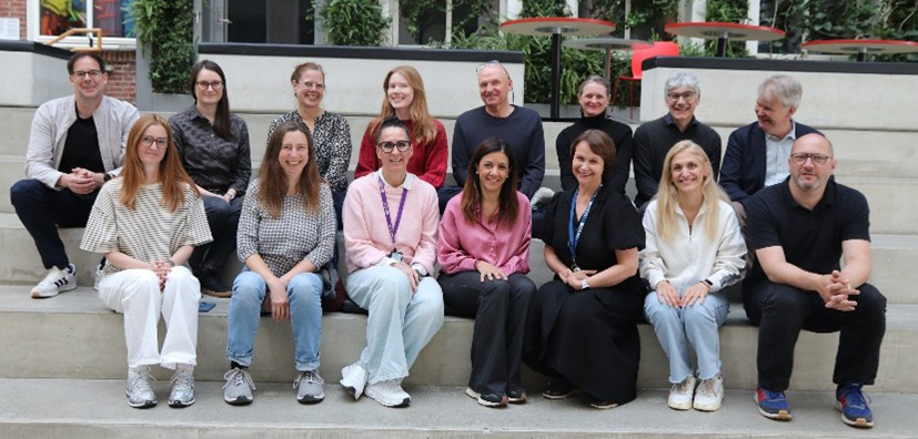 Group photo of several people seated together on indoor steps. The individuals are arranged in two rows and facing the camera, with green plants and tables in the background. Photo