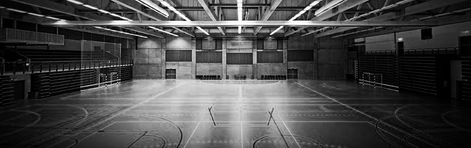 A large, empty indoor sports facility with multiple basketball courts and bleachers on both sides. The ceiling has visible beams and light fixtures. Photo