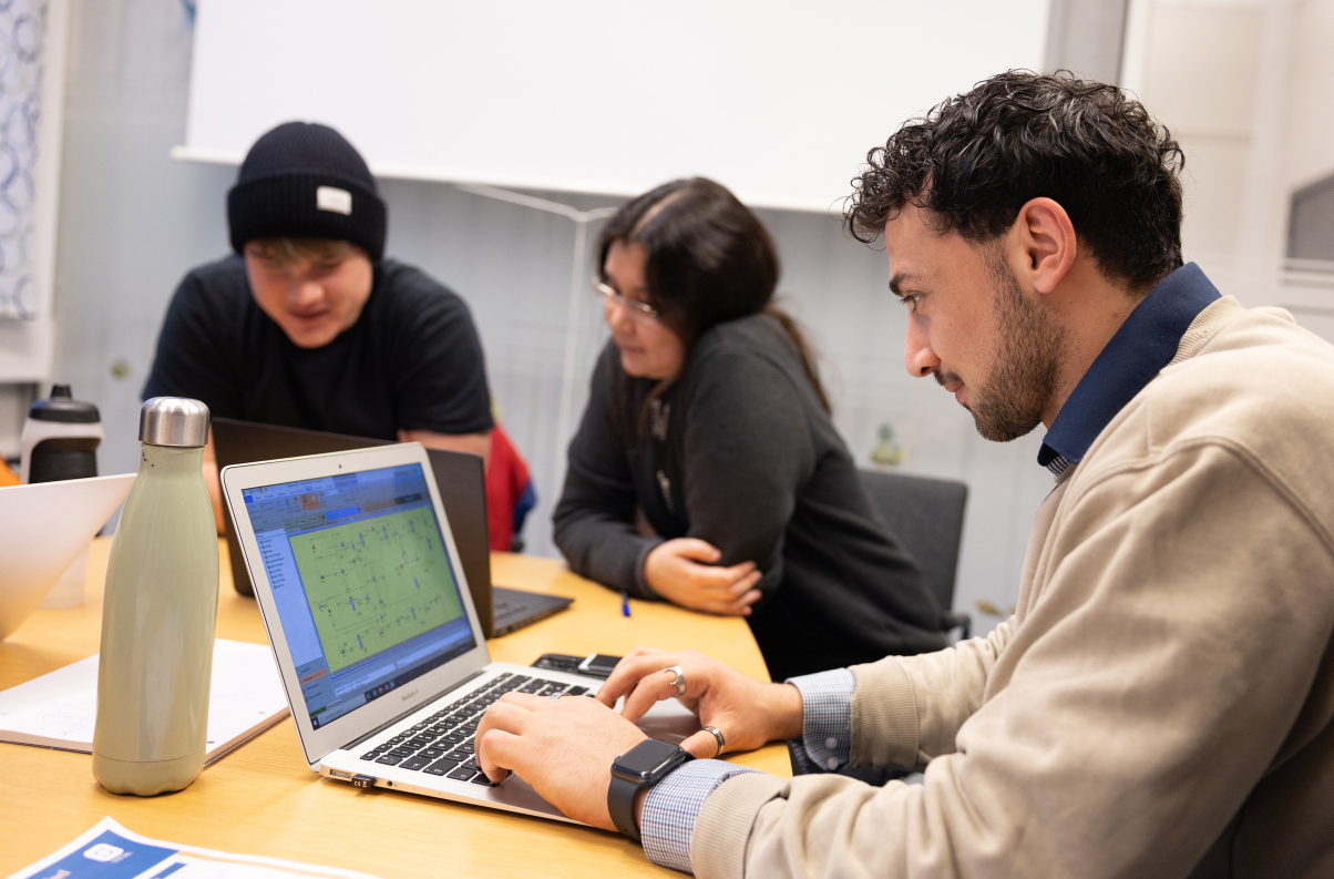 Three students sit around a table. The one closest to the camera smile while looking at his laptop. Photo