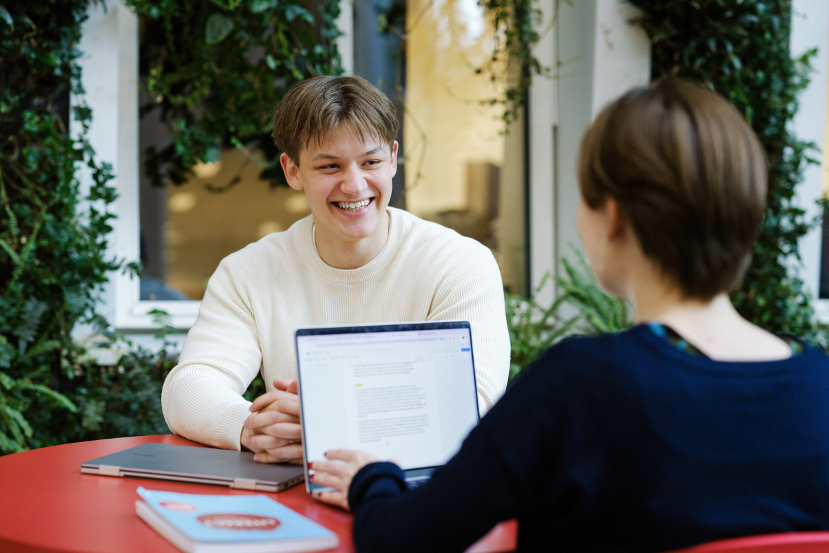 A male student is smiling at another student. Around them is green vegetation. Photo