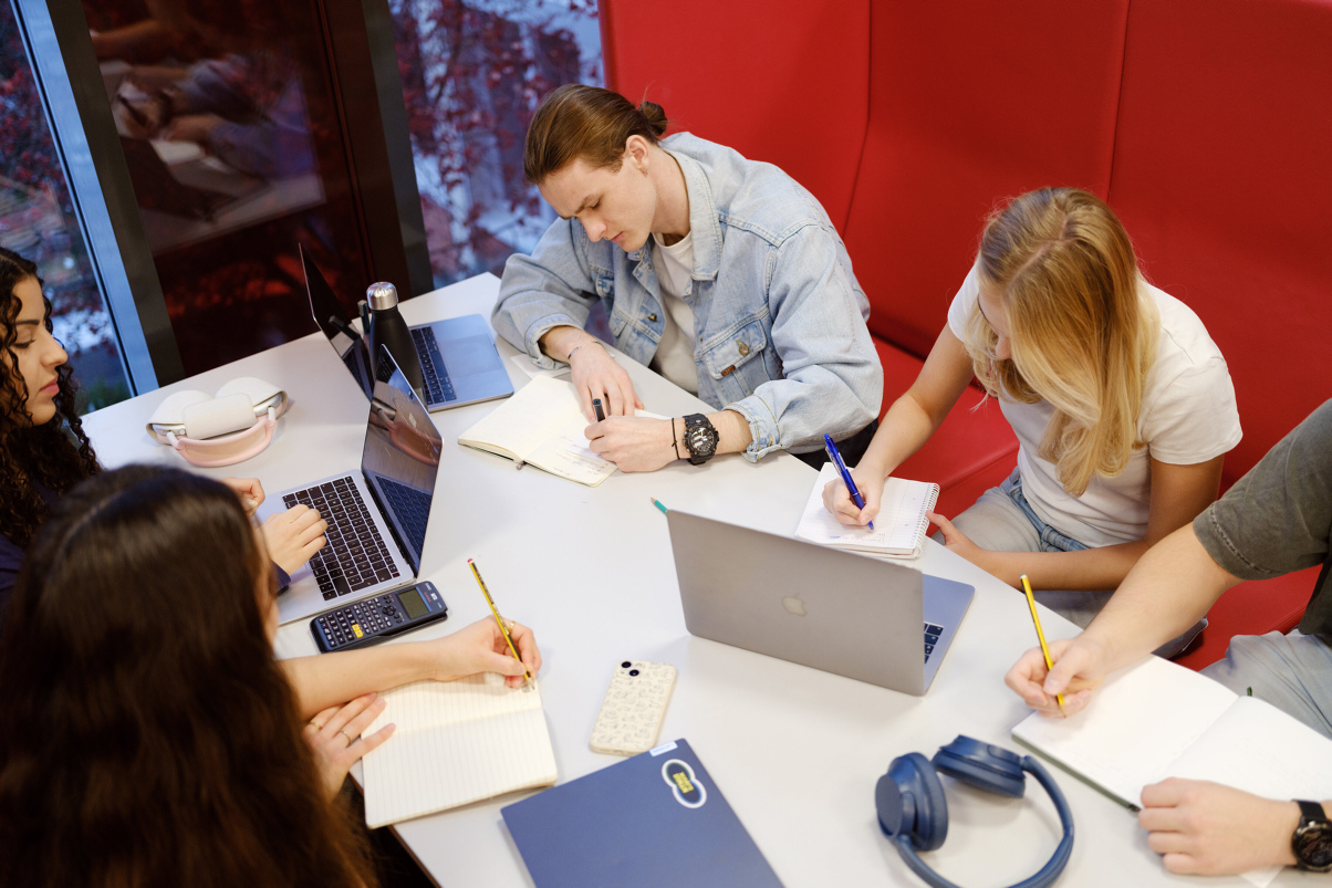 Four students sit writing on notepats with open laptops around a table. Photo