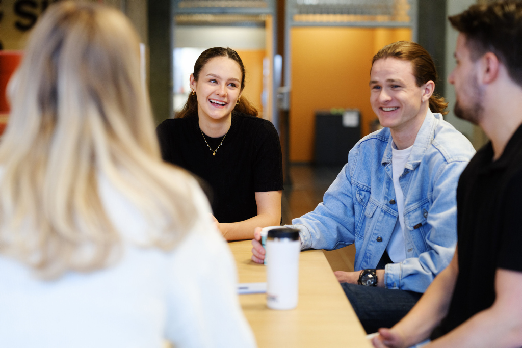 Three students sit around a table smiling and talking. Photo