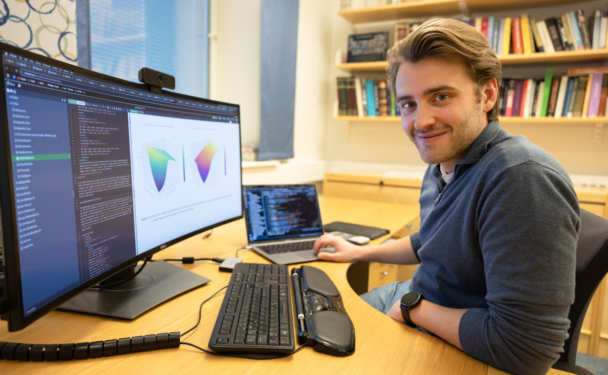 A student is sitting in front of a computer with colourful graphics on the screen. He smiles at the camera. Photo