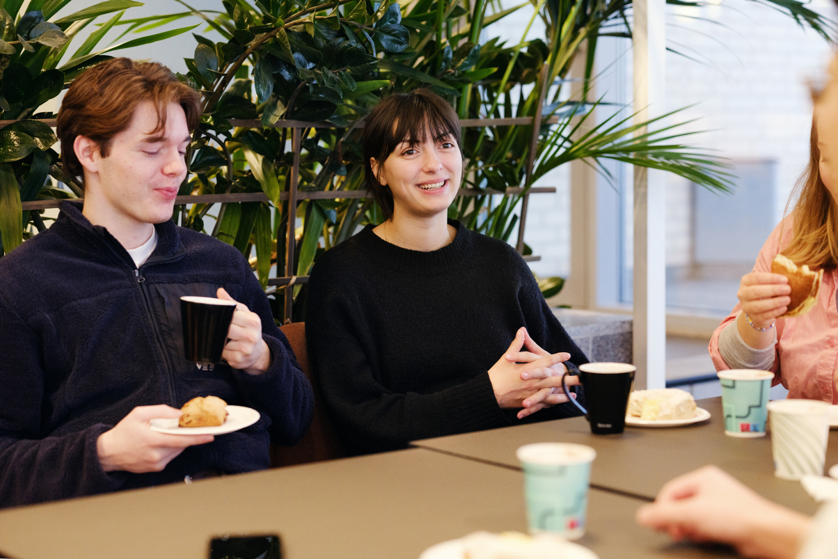 Three students sit in a green environment. One student is in focus smiling. Photo
