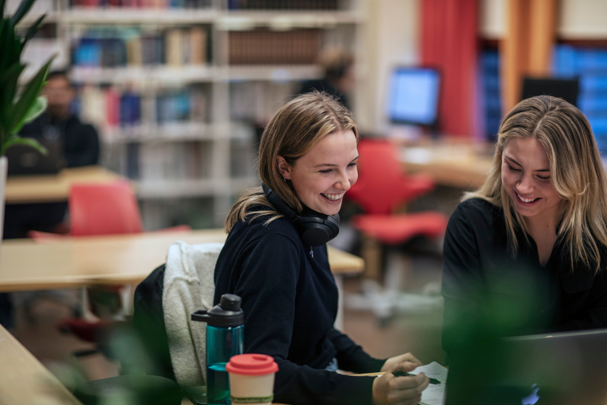 Two female students smiling and looking at a computer screen. In the background we can see books, chairs and desks. 