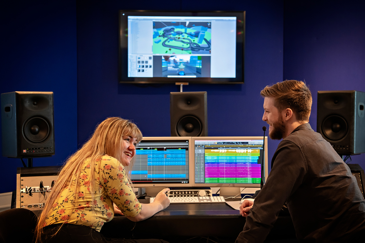 Two people by a mixing desk in a studio, smiling at each other