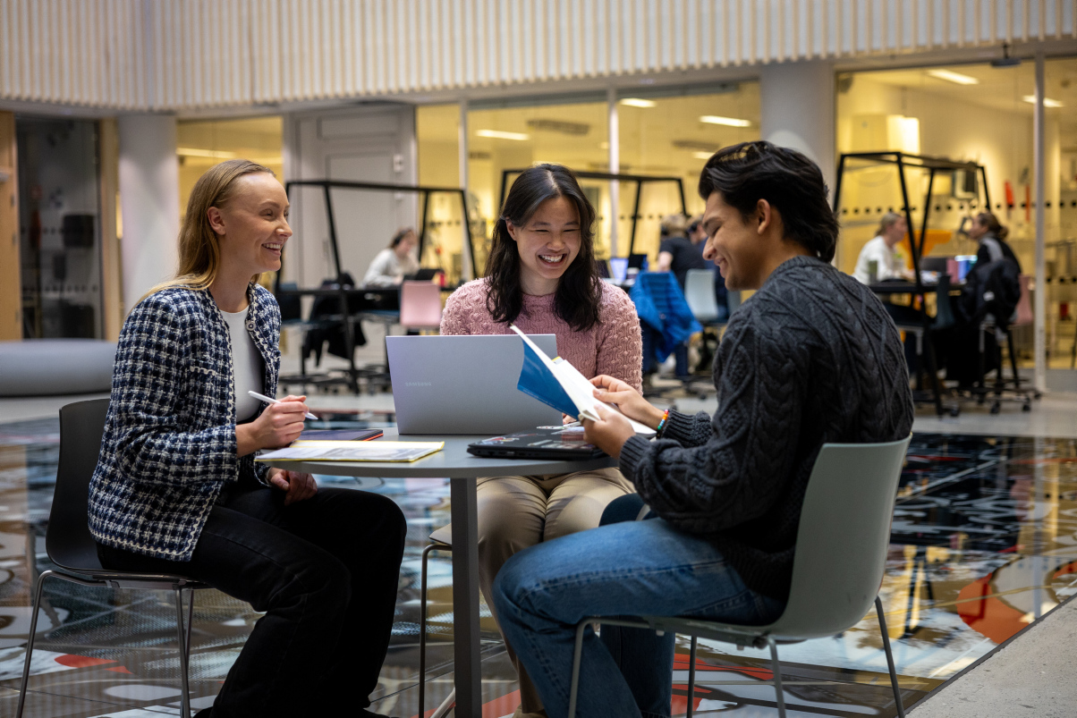 Three students are sitting around a table with books and laptops talking and smiling. Photo