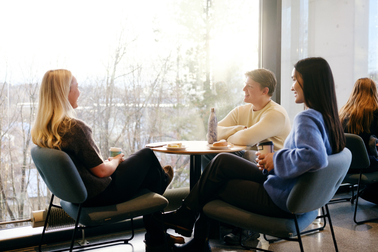 Three students sit around a table talking and smiling. Behind them is a large window where you can see many trees. Photo