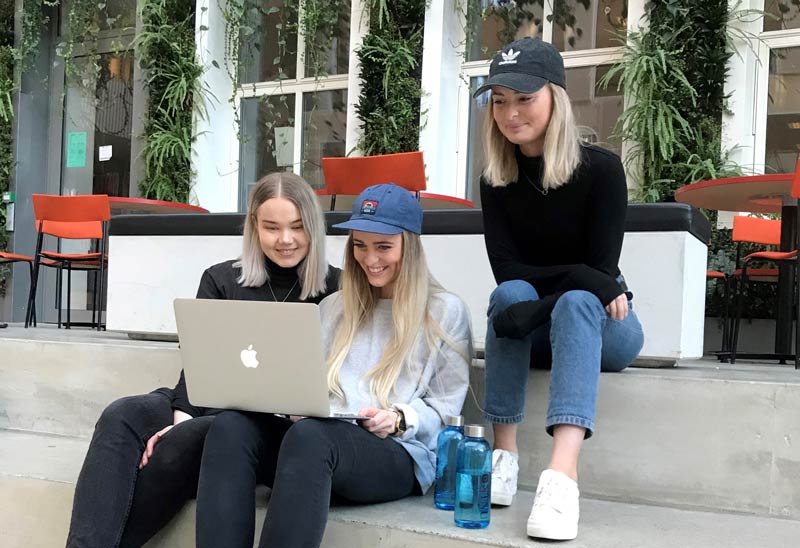 Three students sitting in a staircase looking at a computer. Photo.
