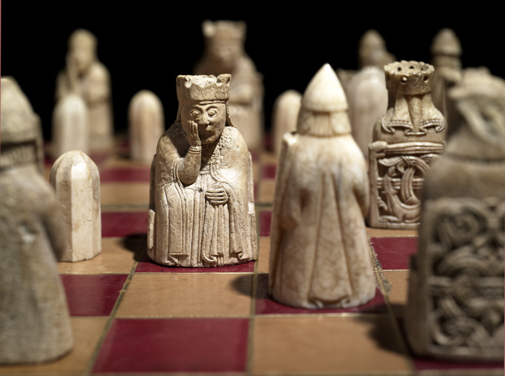A yellow and red chess board with several pieces carved from sea ivory. All the pieces but one queen have their backs turned, showing beautifully carved chair backs. The queen who looks out has one hand pressed to her face and staring eyes.