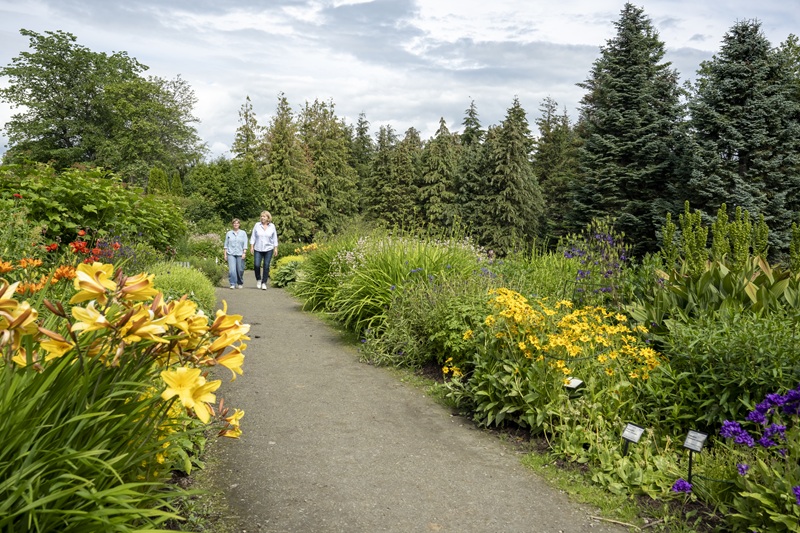 Two women walking along a gravel path flanked by colorful flower beds.