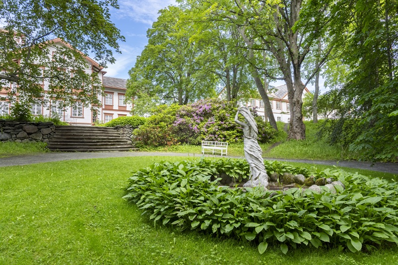 Statue of a woman surrounded by flower beds, with a historic manor house in the background.